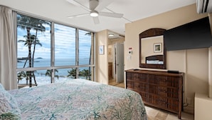 A bedroom with a large bed, a ceiling fan, a TV, and a view of the ocean. - Kamaole Nalu, #405 2 Bedroom Condo (Kihei)