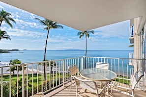 A balcony with a glass table and chairs, overlooking a beach and ocean. - Kamaole Nalu, #405 2 Bedroom Condo (Kihei)
