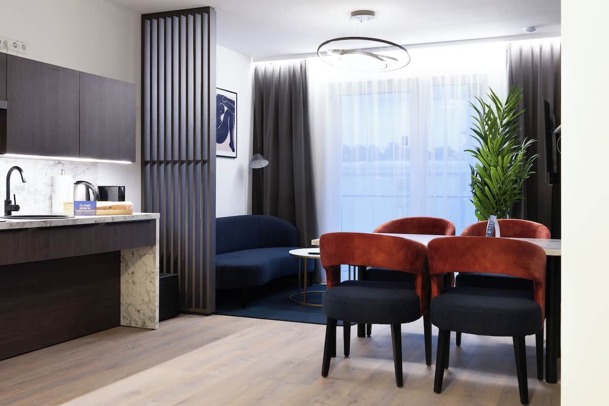 Een moderne hotelkamer met een kitchenette, een eettafel met stoelen, een bank en een groot raam met gordijnen.