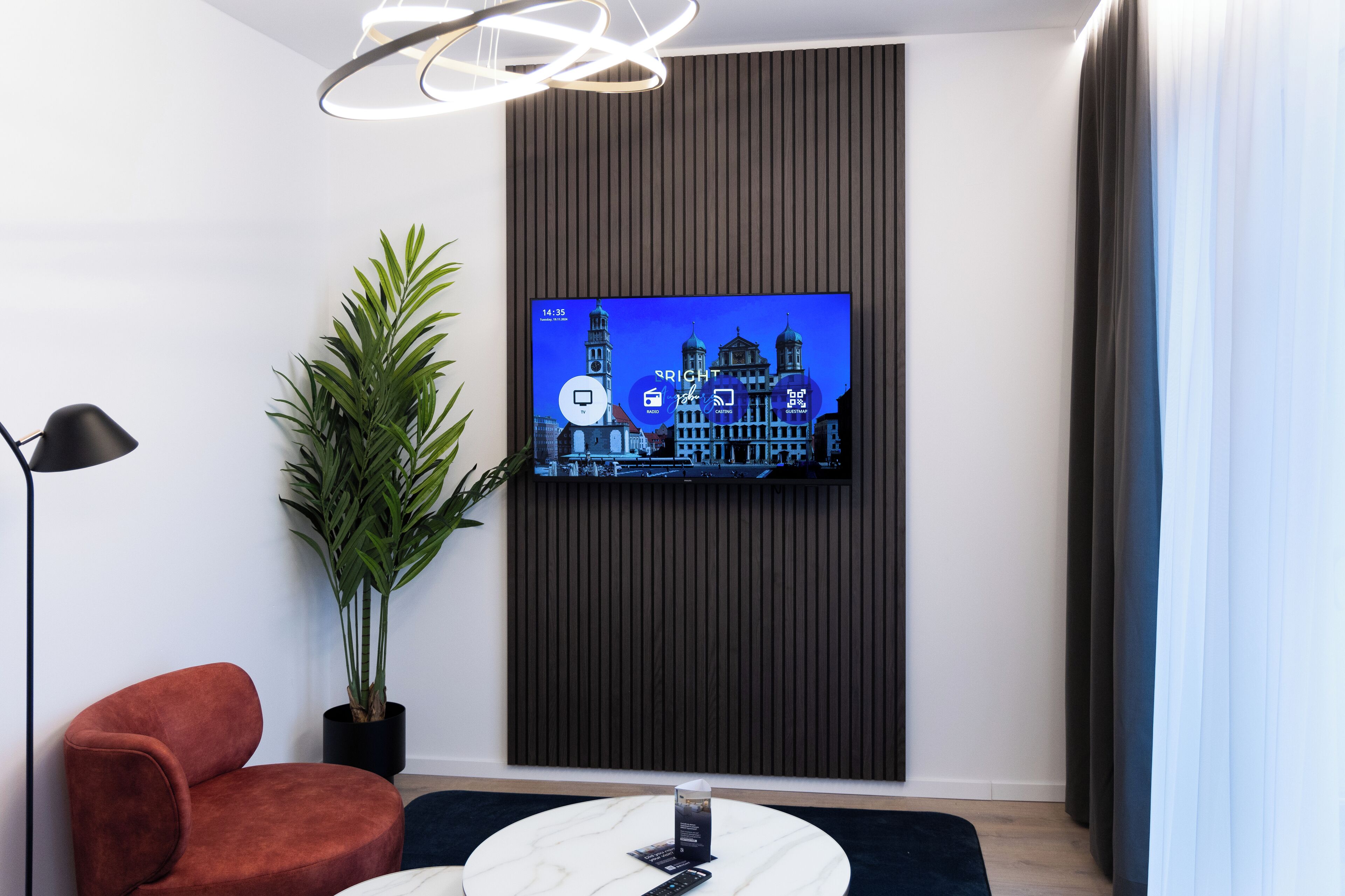 A modern room with a TV mounted on a dark panel, a red chair, a potted plant, and a round table with a remote control.
