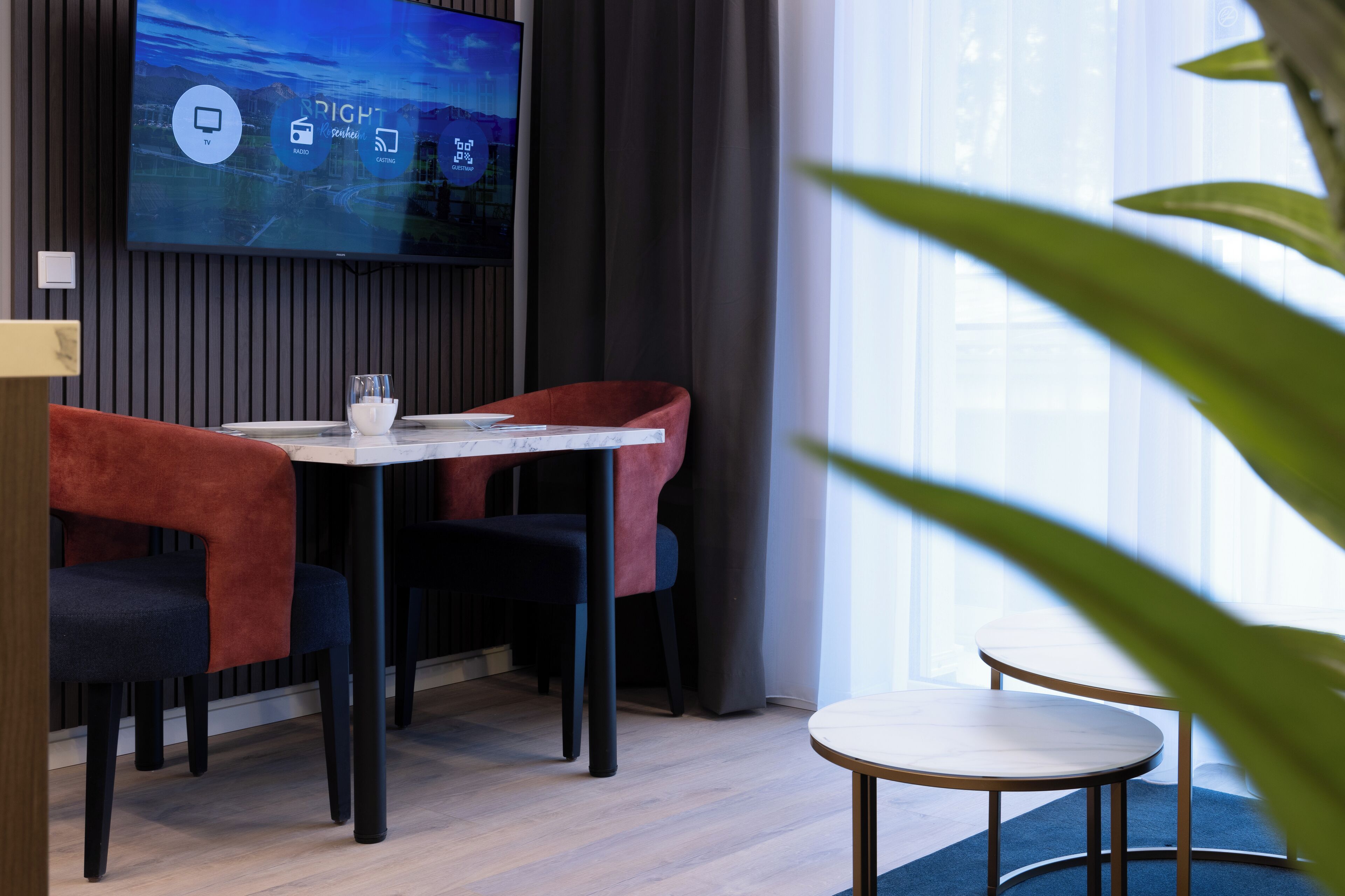 Ett modernt rum med en TV som hänger på väggen och visar en meny, ett bord med stolar och en växt i förgrunden.