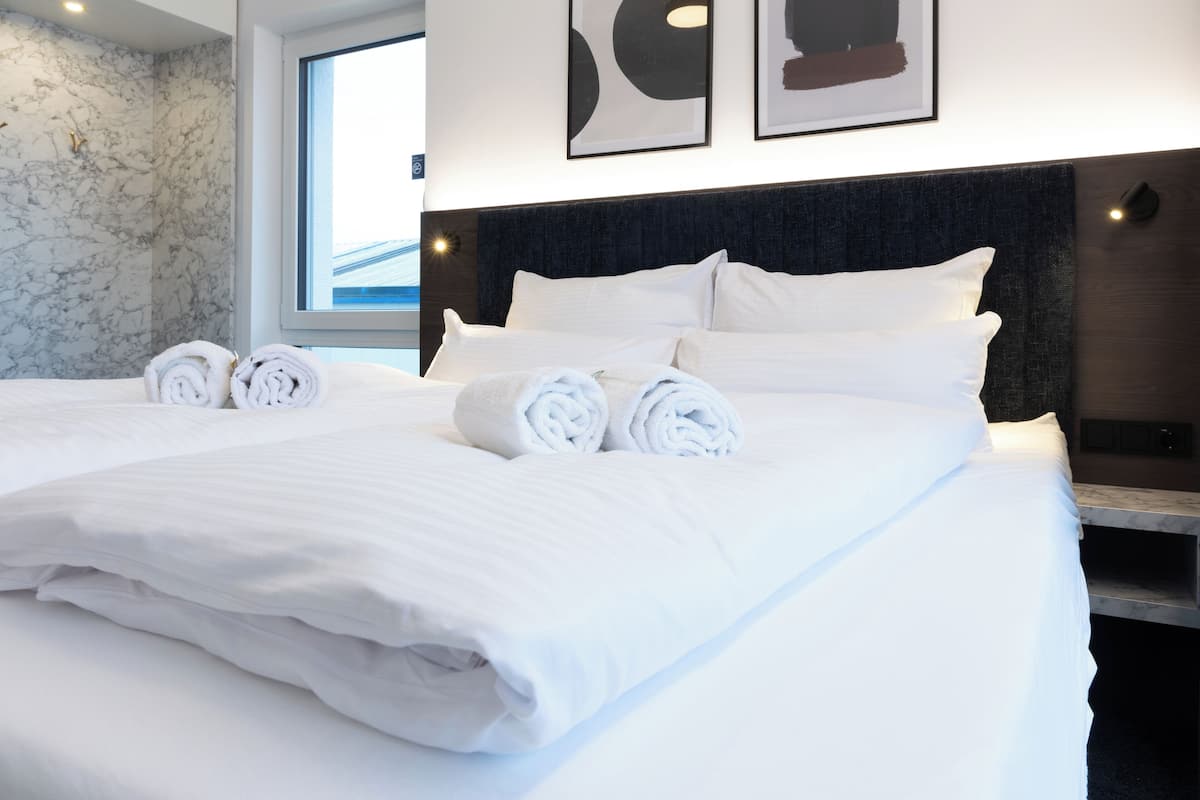 Een net opgemaakt bed met witte linnens en opgerolde handdoeken, een donker hoofdbord en kunstwerken in een lijst aan de muur.