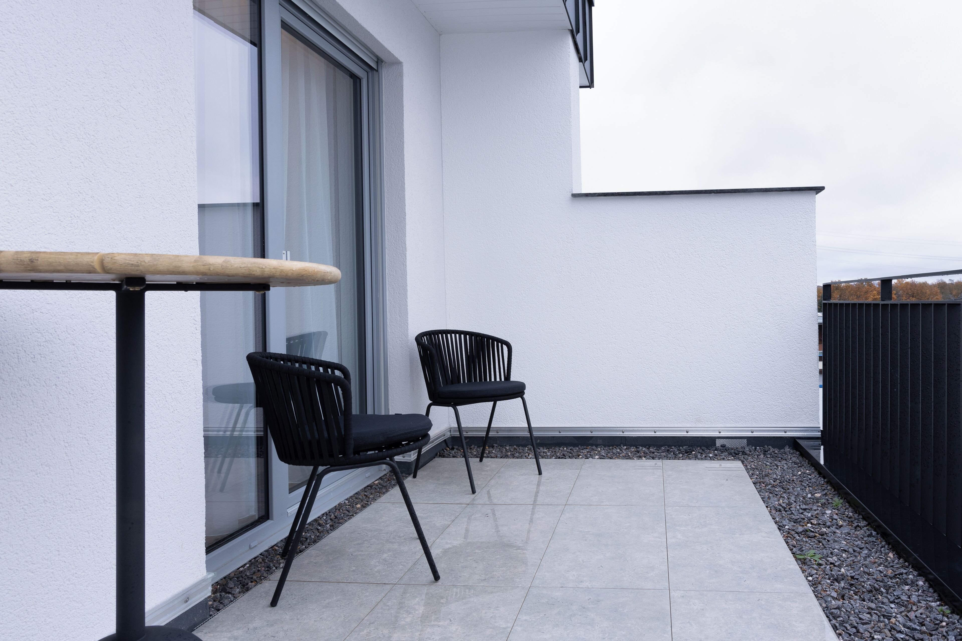 Ein Balkon mit zwei schwarzen Stühlen, einer Holzbank und einer weißen Wand.