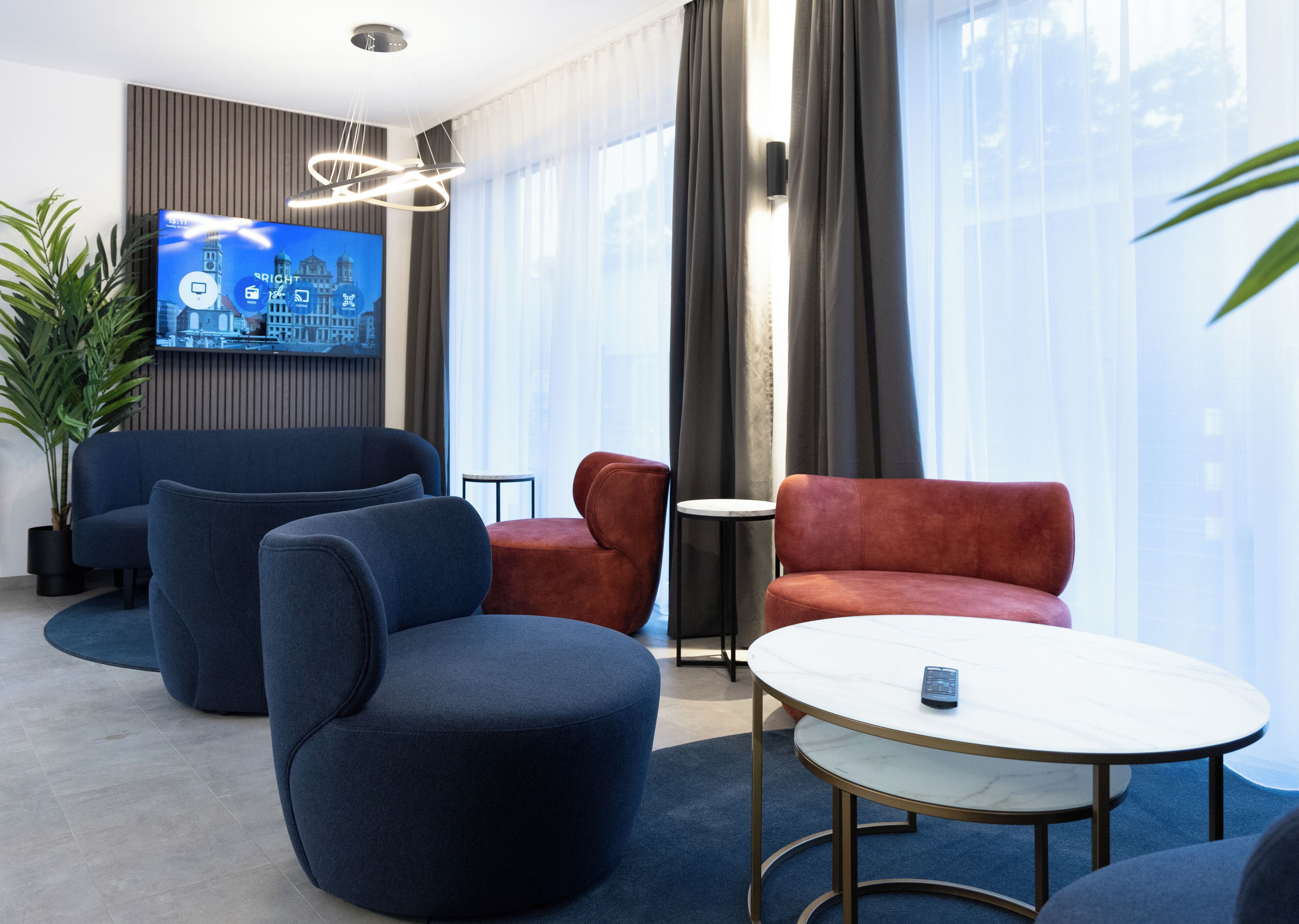 En moderne hotellobby med et stort fladskærms-tv, behagelige siddepladser, et lille rundt bord og en potteplante.