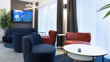 En moderne hotellobby med et stort fladskærms-tv, behagelige siddepladser, et lille rundt bord og en potteplante.