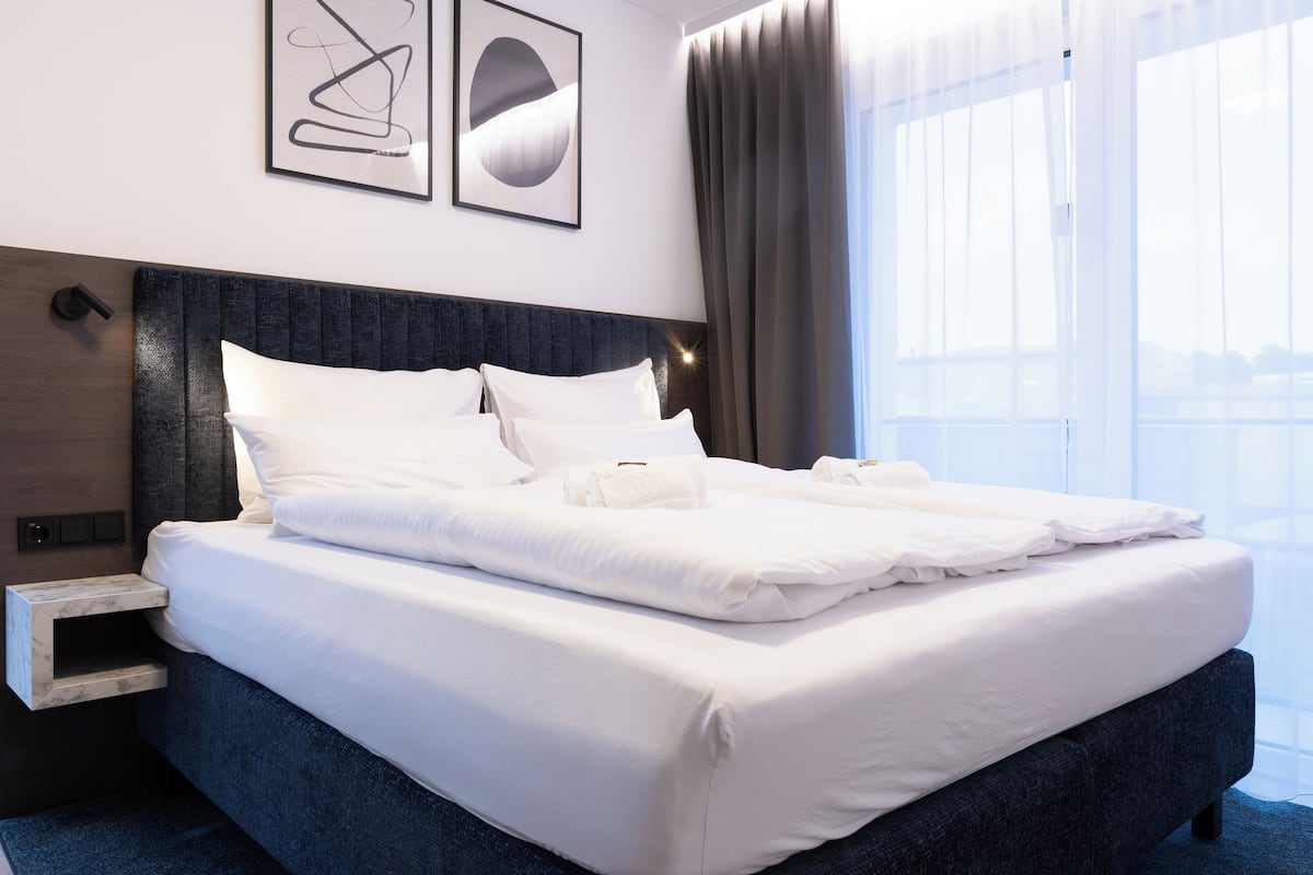 Een net opgemaakt bed in een hotelkamer, met een donker hoofdbord, een nachtkastje en een groot raam met gordijnen.
