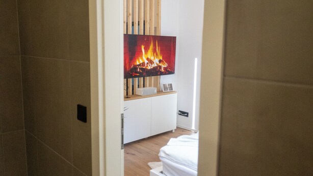 A modern bathroom with a shower, a toilet, and a sink. A television is mounted on the wall displaying a fire. - DLG-STAYS - "Pure 23" (Iserlohn)