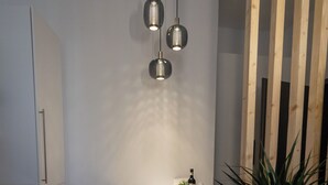 A modern kitchen with white cabinetry, a dining table with a chair, pendant lighting, and a potted plant. - DLG-STAYS - "Pure 23" (Iserlohn)