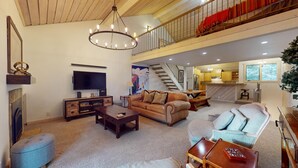 A spacious living area with a large TV, a wooden entertainment unit, a sofa, a coffee table, and a staircase leading to an upper level.