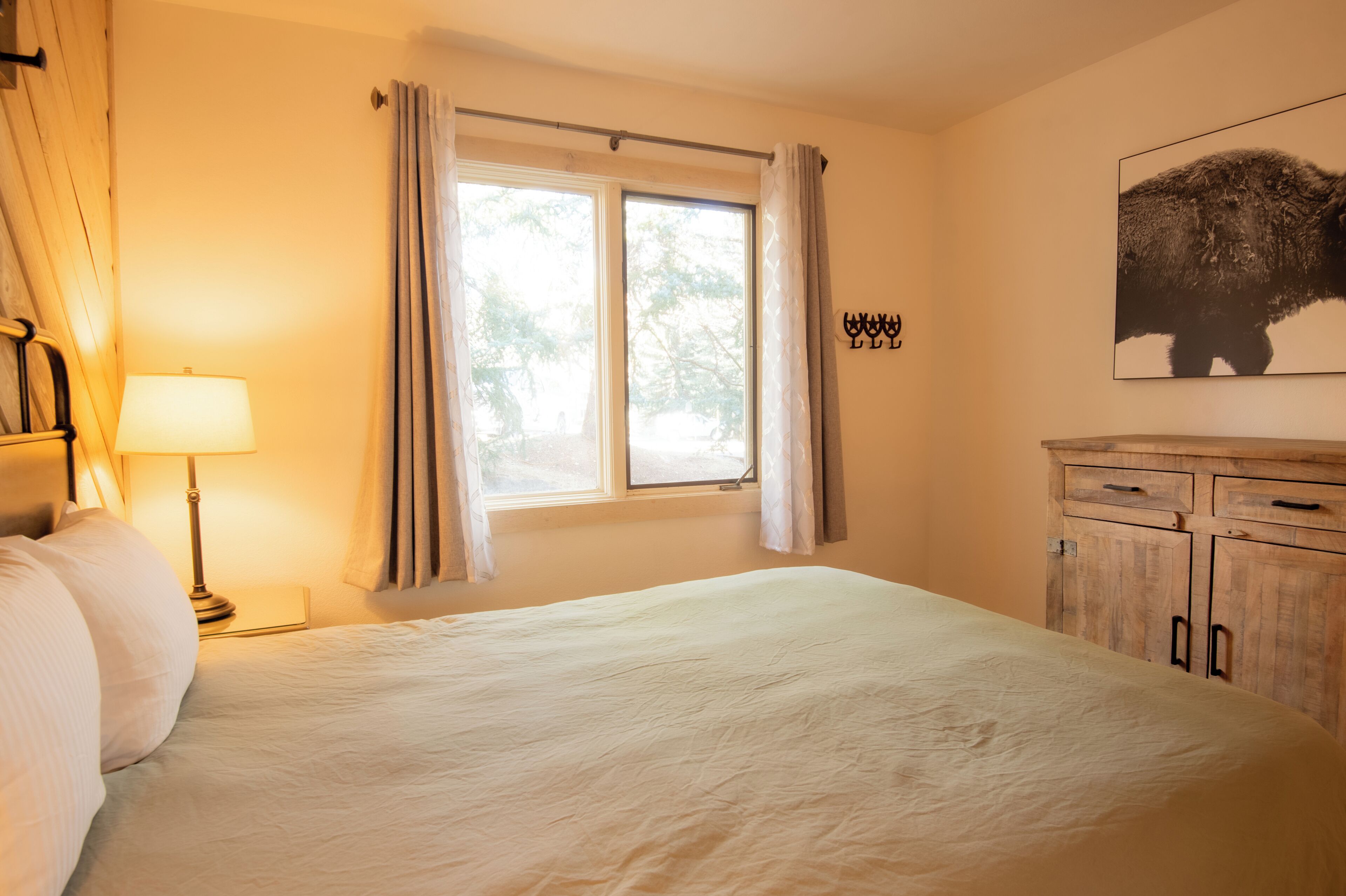 Quarto com cama grande, janela com cortinas, luminária, guarda-roupa de madeira e quadro de um búfalo fixado na parede.