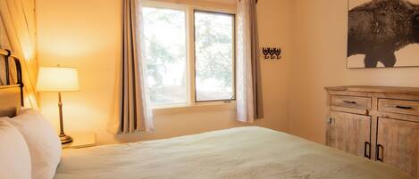 Quarto com cama grande, janela com cortinas, luminária, guarda-roupa de madeira e quadro de um búfalo fixado na parede.
