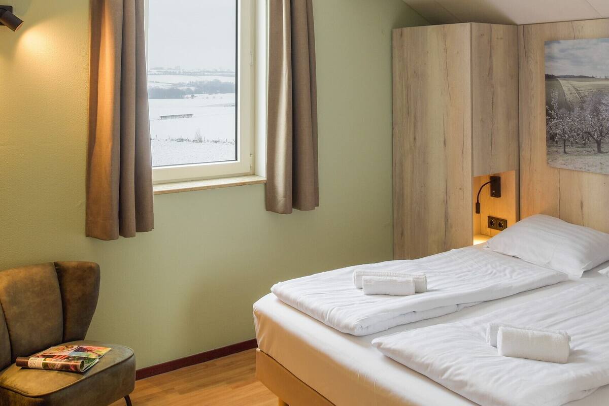 A hotel room with a bed, a chair, a window with curtains, a painting, and a lamp.