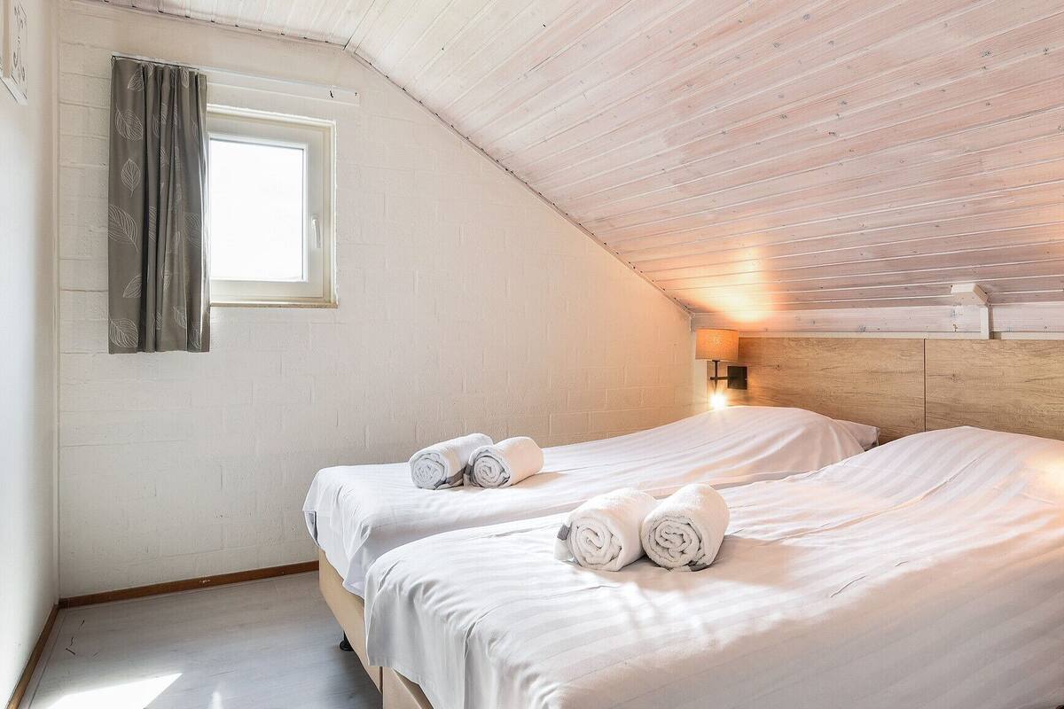 A cozy, well-lit attic bedroom with two beds, a window with curtains, and a wooden ceiling.