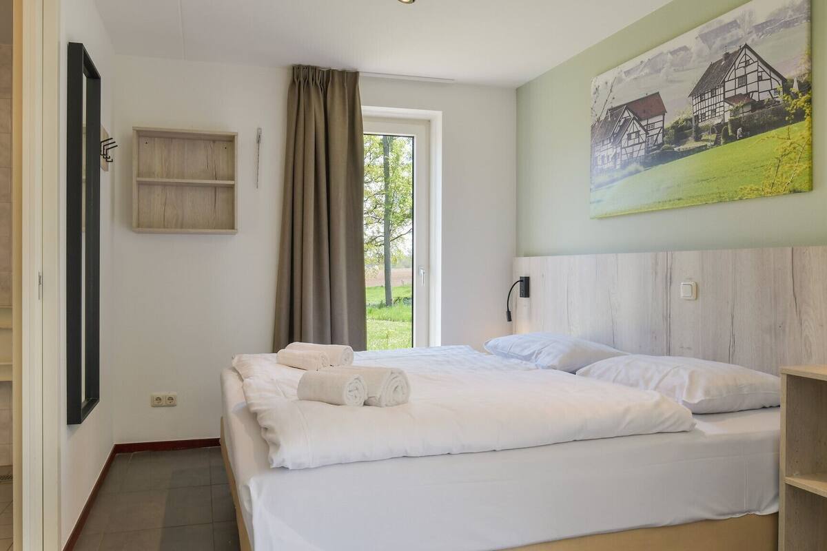 A hotel room with a bed, a shower, a window with curtains, and a painting on the wall.