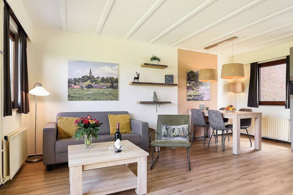 A living room with a sofa, a coffee table with a bottle and flowers, a dining area with chairs and a table, and a kitchen with appliances.