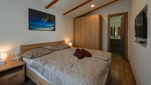 Une chambre à coucher comprenant un lit, une tête de lit en bois, des tables de chevet, un tableau représentant une plage, une armoire et un téléviseur fixé au mur.