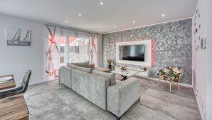 A modern living room with a grey sectional sofa, a flat-screen TV mounted on the wall, and a dining area with a wooden table and chairs. - Peaceful Blooming Villa With Pool (Rovinj)
