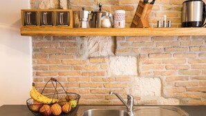 Une cuisine avec un mur en briques, une étagère en bois où sont rangés des ustensiles de cuisine, un panier de fruits et un évier en acier inoxydable.