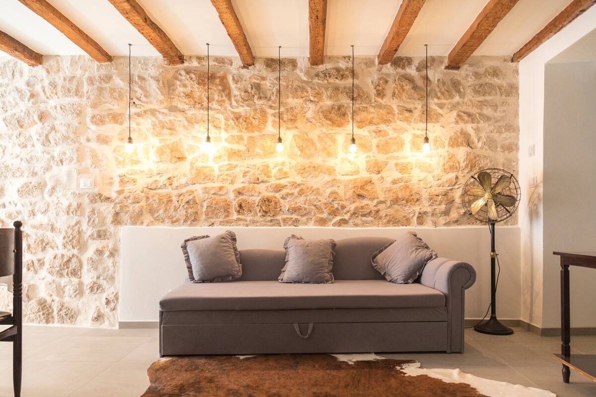 A cozy living room with a stone wall, a grey sofa with cushions, a wooden floor, and a fan.