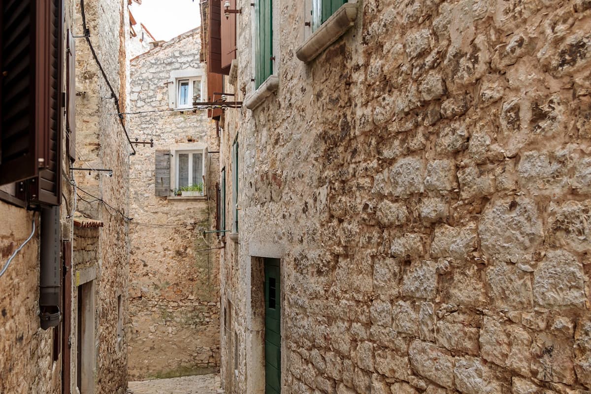 A narrow stone alleyway with traditional architecture, including wooden shutters and a small window with plants.