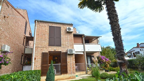 Uma casa de dois andares em tijolo, com varanda, aparelhos de ar condicionado e um jardim bem cuidado com diversas plantas e flores.