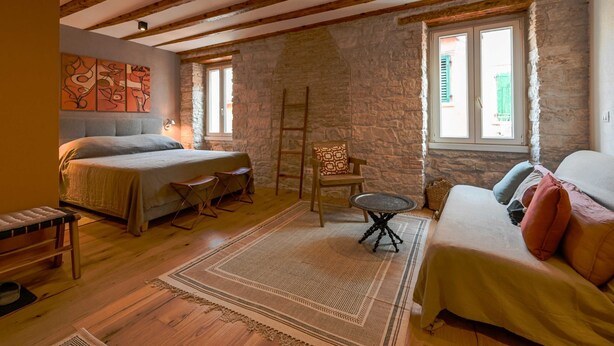 A bedroom with a bed, a chair, a small table, and a sofa. There are stone walls and wooden beams on the ceiling. - Alluring Studio Apartment Downtown Rovinj (Rovinj)