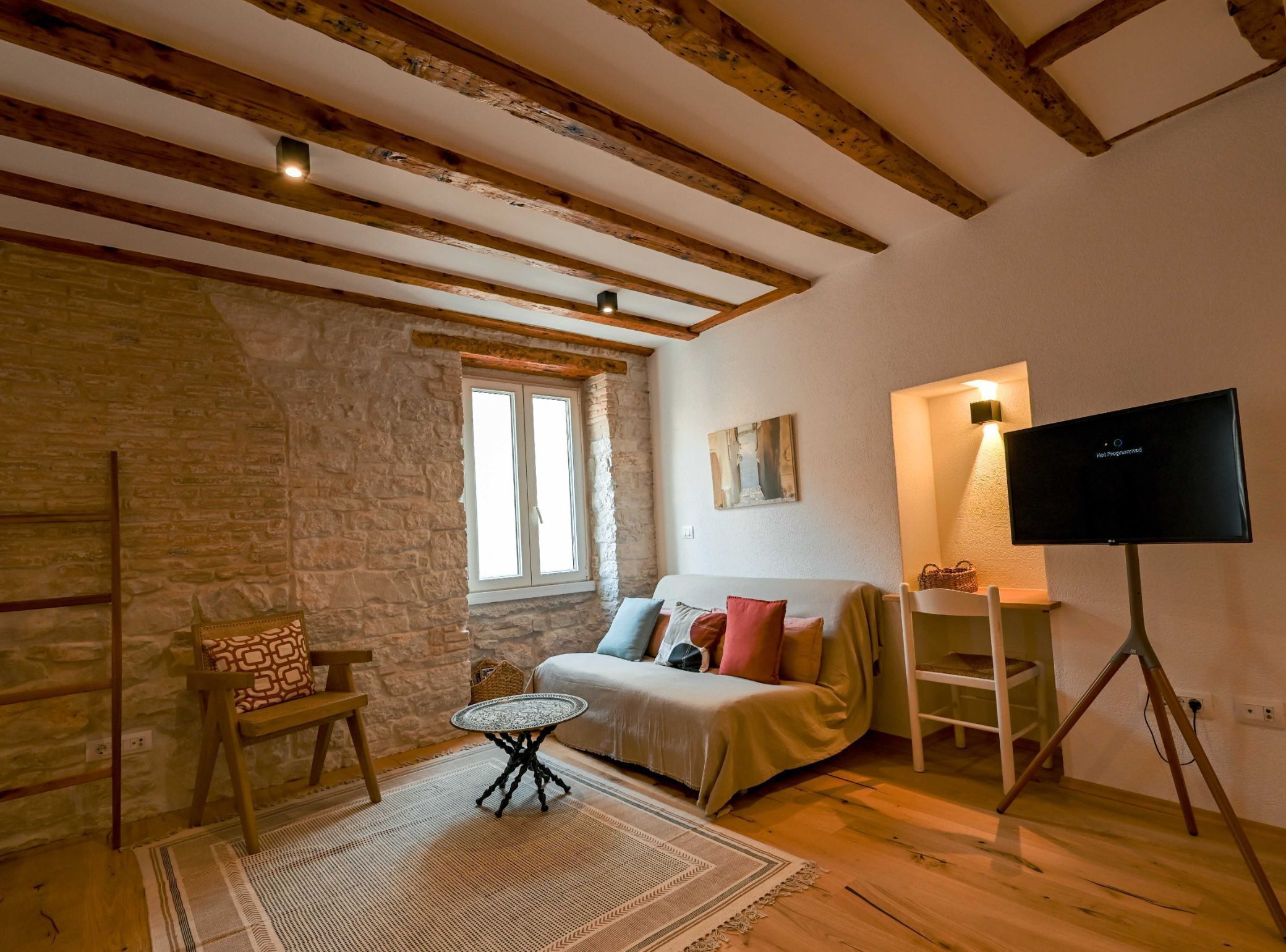 A cozy room with exposed wooden beams, a stone wall, a sofa with cushions, a small round table, and a television on a tripod stand.