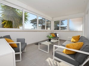 A sunlit patio with grey sofas and white tables, large windows, and a view of trees and sky. - Tranquil and Bright Apartment With Loggia (Rovinj)