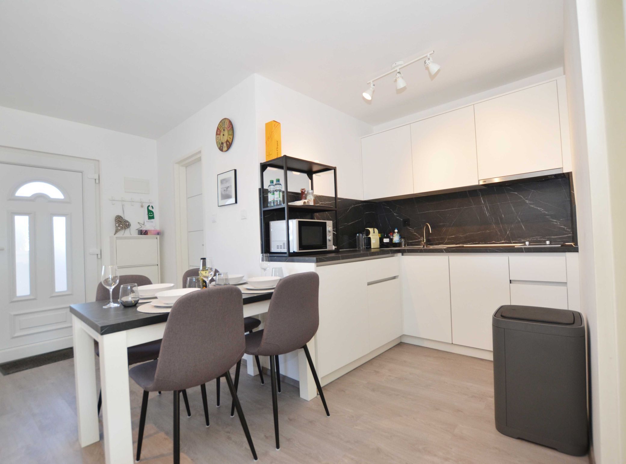 A modern kitchen with white cabinets, a black countertop, and a microwave. There is a dining table with chairs and a trash bin.