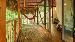 A wooden deck with a hammock, bamboo fence, and a glass door leading to a bedroom with a bed and curtains. - Cozy Cabin Nestled in Bamboo Forest (El Caimo)