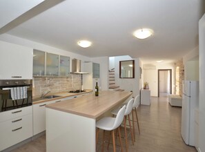 A modern kitchen with white cabinetry, a central island with bar stools, a built-in oven, and a stone accent wall. - Forest Breeze Retreat in Zminj (Sveti Petar u Sumi)