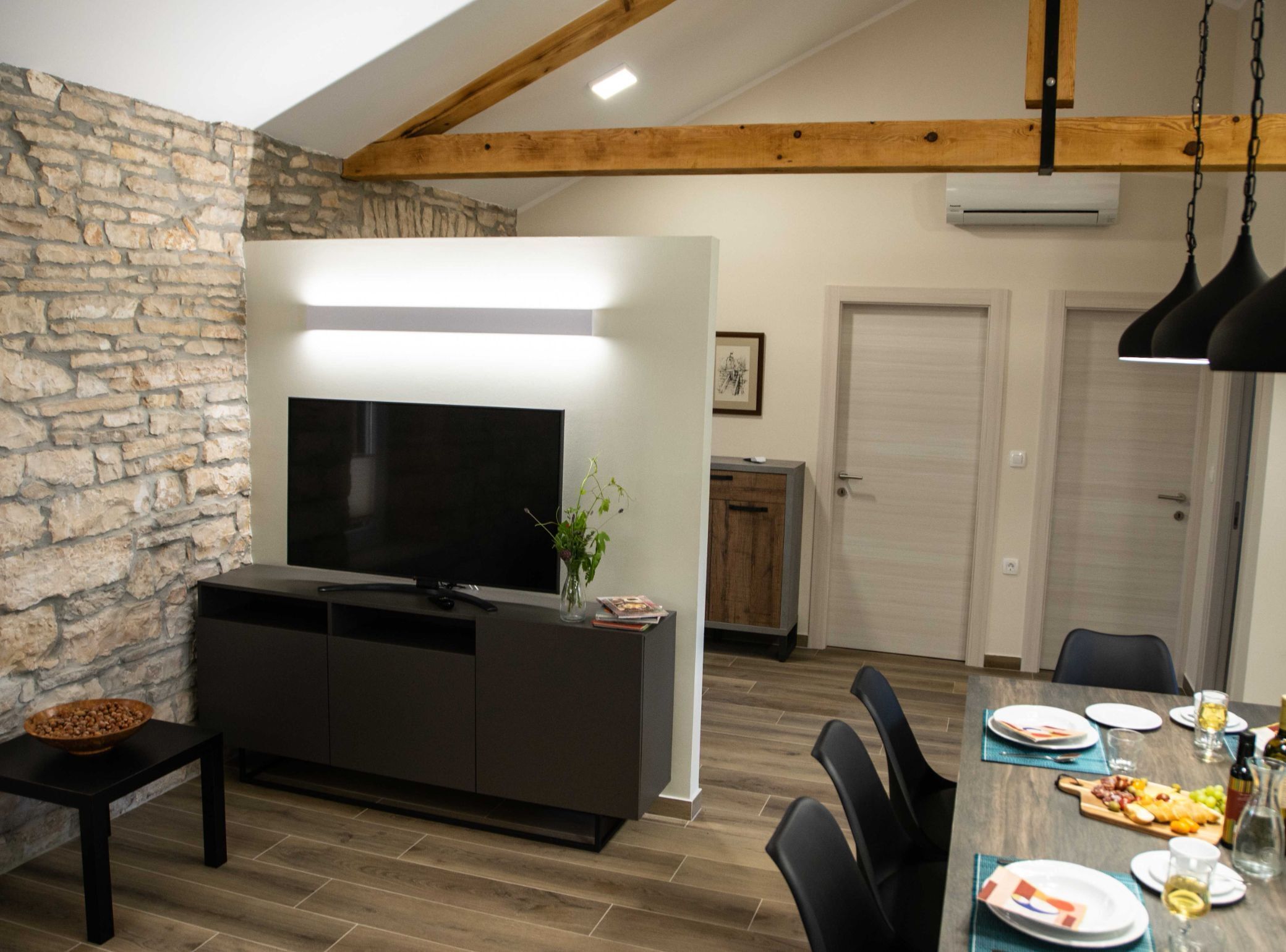 A modern living room with a stone wall, a flat-screen TV, a dark wooden entertainment unit, and a dining area with a wooden table set for a meal.