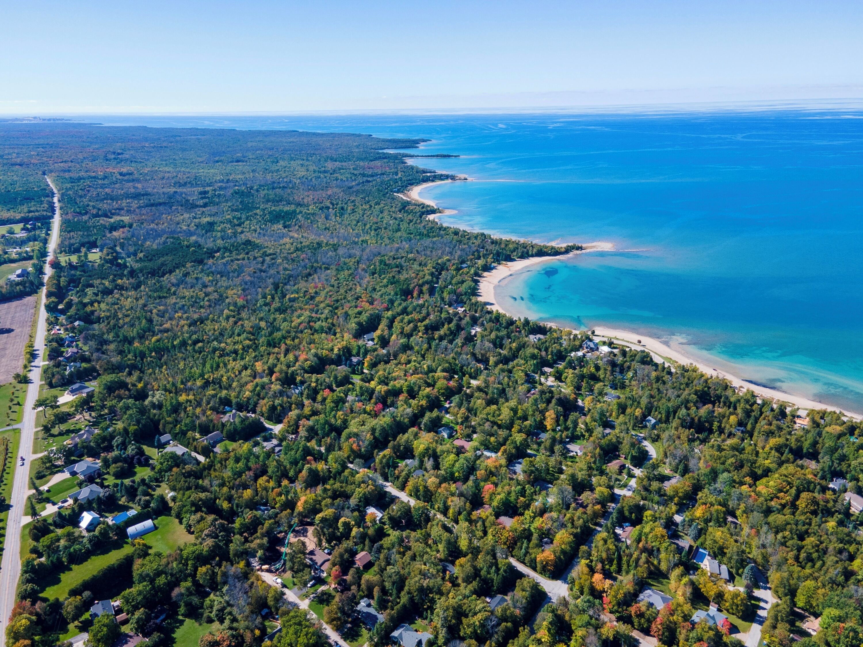 An aerial view of a coastal residential area with dense forestation, a curving beach, and a clear blue sea.