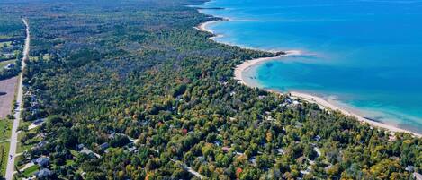 An aerial view of a coastal residential area with dense forestation, a curving beach, and a clear blue sea.