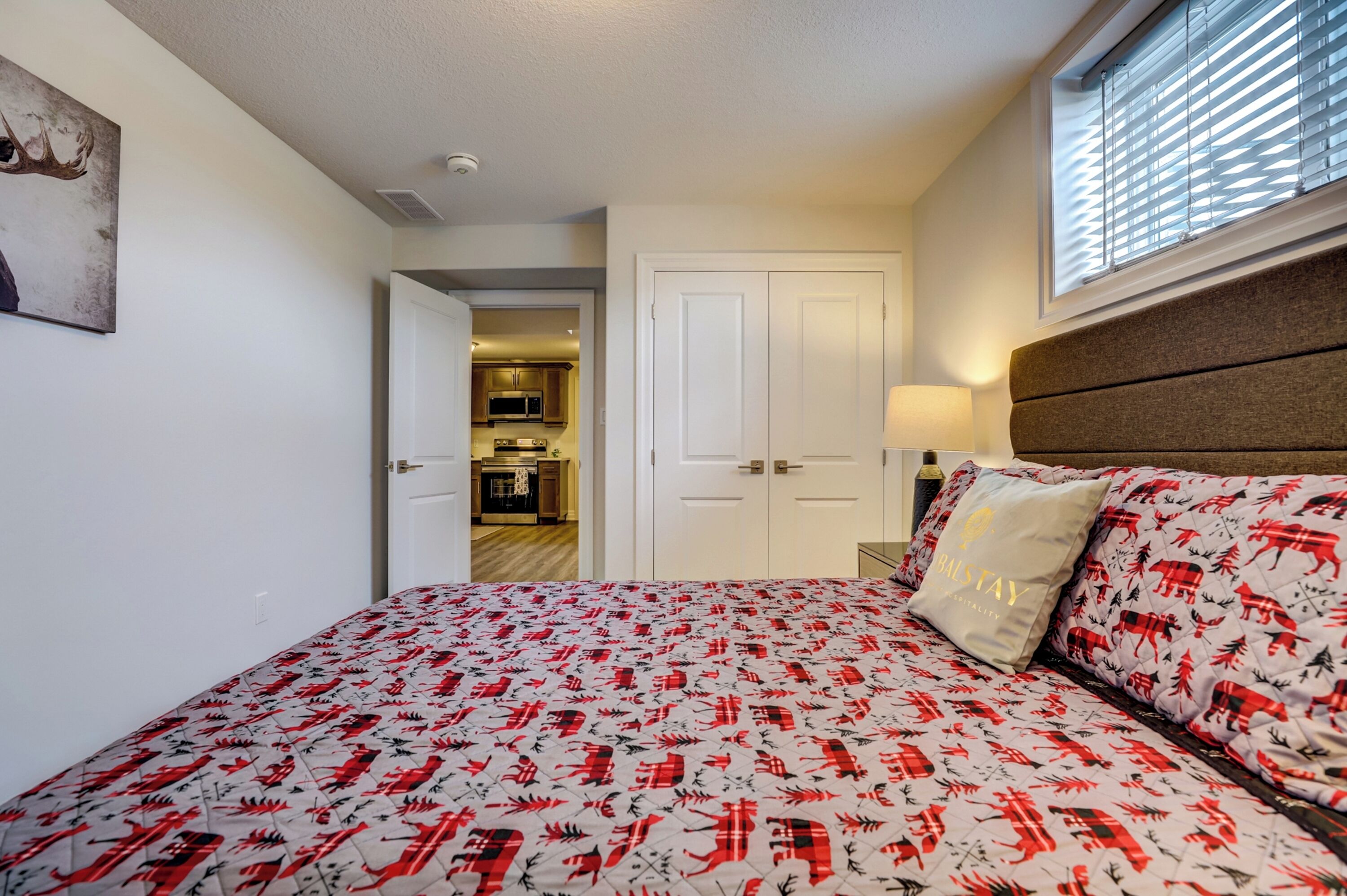 A bedroom with a bed, a nightstand, a lamp, and a door leading to a kitchen.