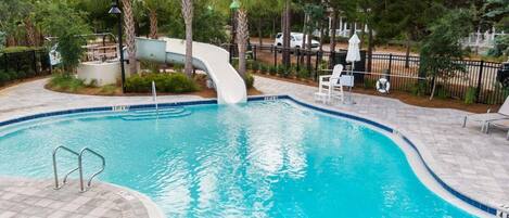 A swimming pool with a slide, surrounded by palm trees and a paved deck with lounge chairs.
