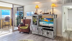 A living room with a television displaying 'OCMD Getaways', a wicker chair, and a view of the ocean through a sliding door. - 20th Floor Ocean Front Condo! Steps to the Beach and Ocean! Atlantis #2004 (Ocean City)