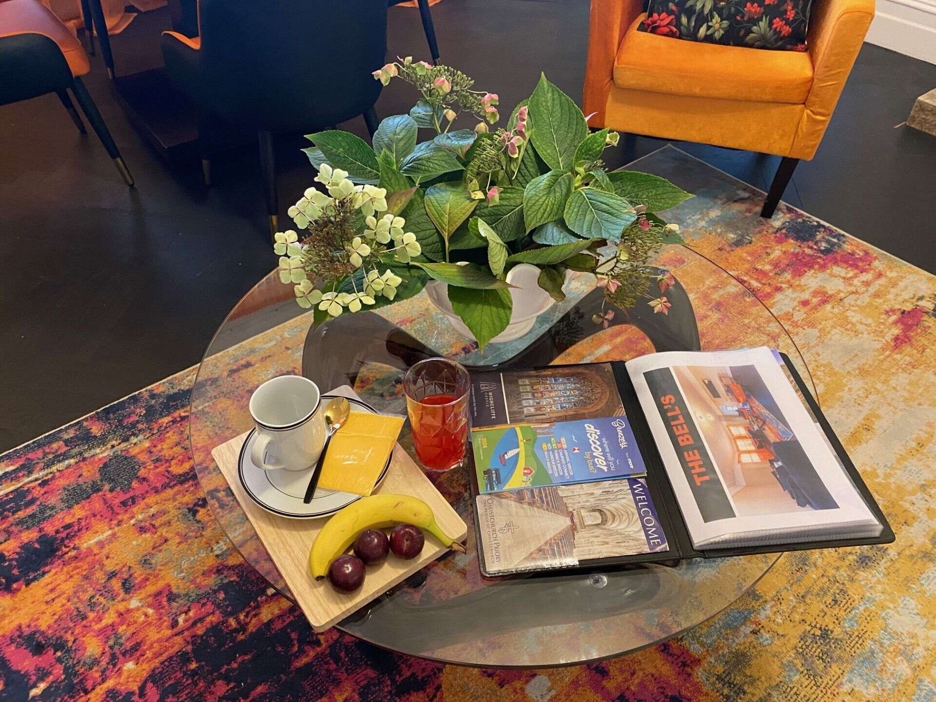 A table with a glass top, a book, a glass of tea, a banana, and a plate with a cup and a slice of cheese.