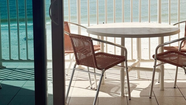 A balcony with a round table and wicker chairs, overlooking a beach and the ocean.