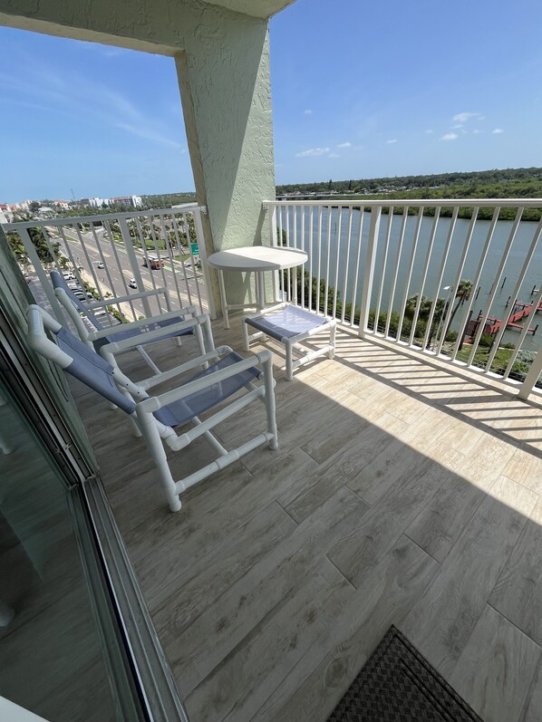A balcony with a view of a road and a body of water. There are two lounge chairs and a small table.