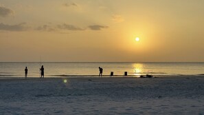 A beach scene with the sun setting over the ocean, silhouettes of people and a chair on the sand. - Holiday Villas III-708 Intracoastal view! Low rates all 2026! (Indian Shores)