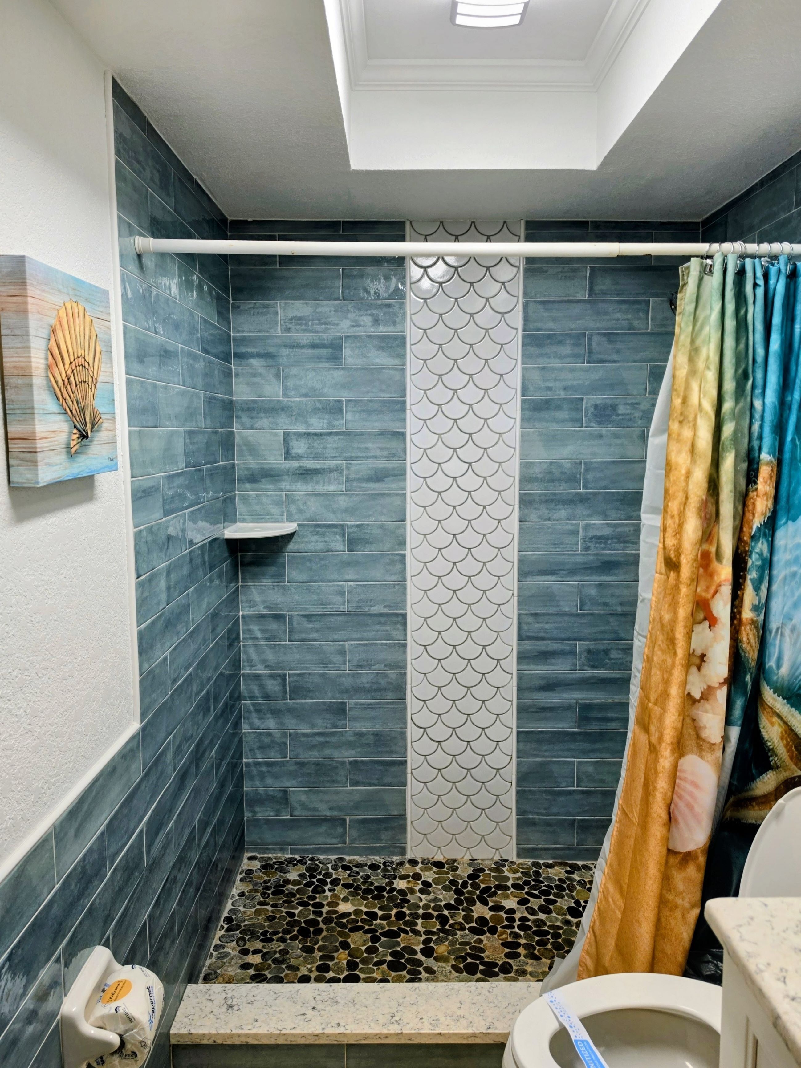 Un área de ducha con azulejos azules, un panel decorativo blanco y una cortina de ducha con estampado de peces.