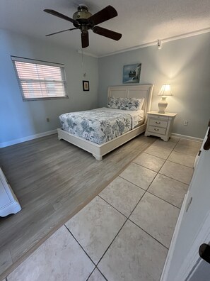 A bedroom with a bed, a nightstand, a ceiling fan, a window with blinds, and a painting on the wall.