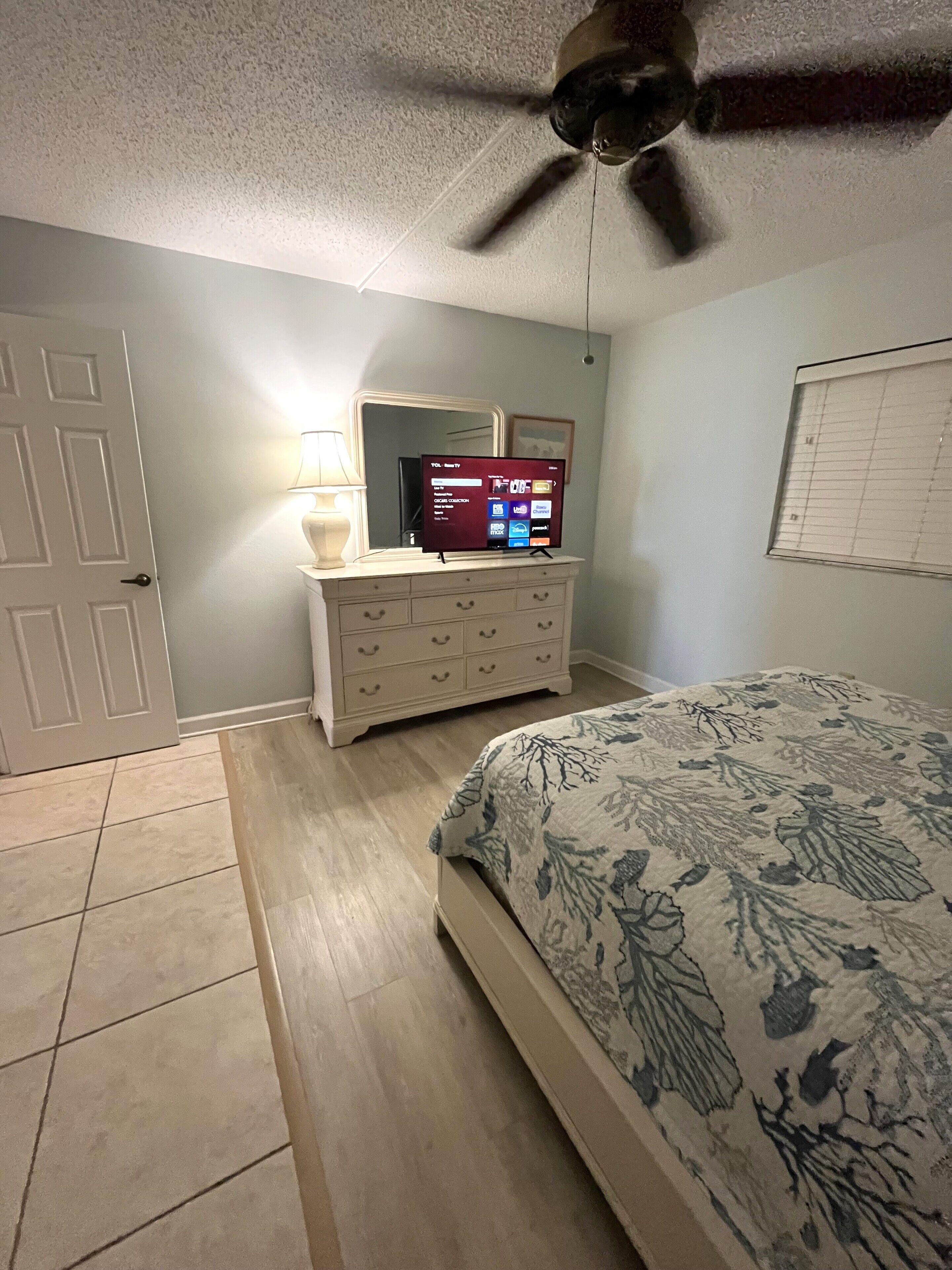 Un dormitorio con cama, cómoda, ventilador de techo, televisor y ventana con persianas.