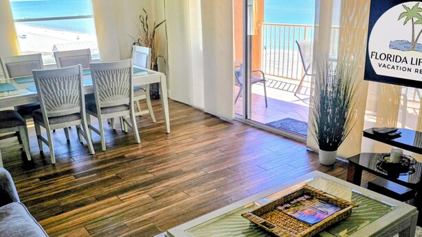 A spacious living room with a wooden floor, a dining area with white chairs, a large window overlooking the beach, and a sign indicating a vacation rental in Florida.