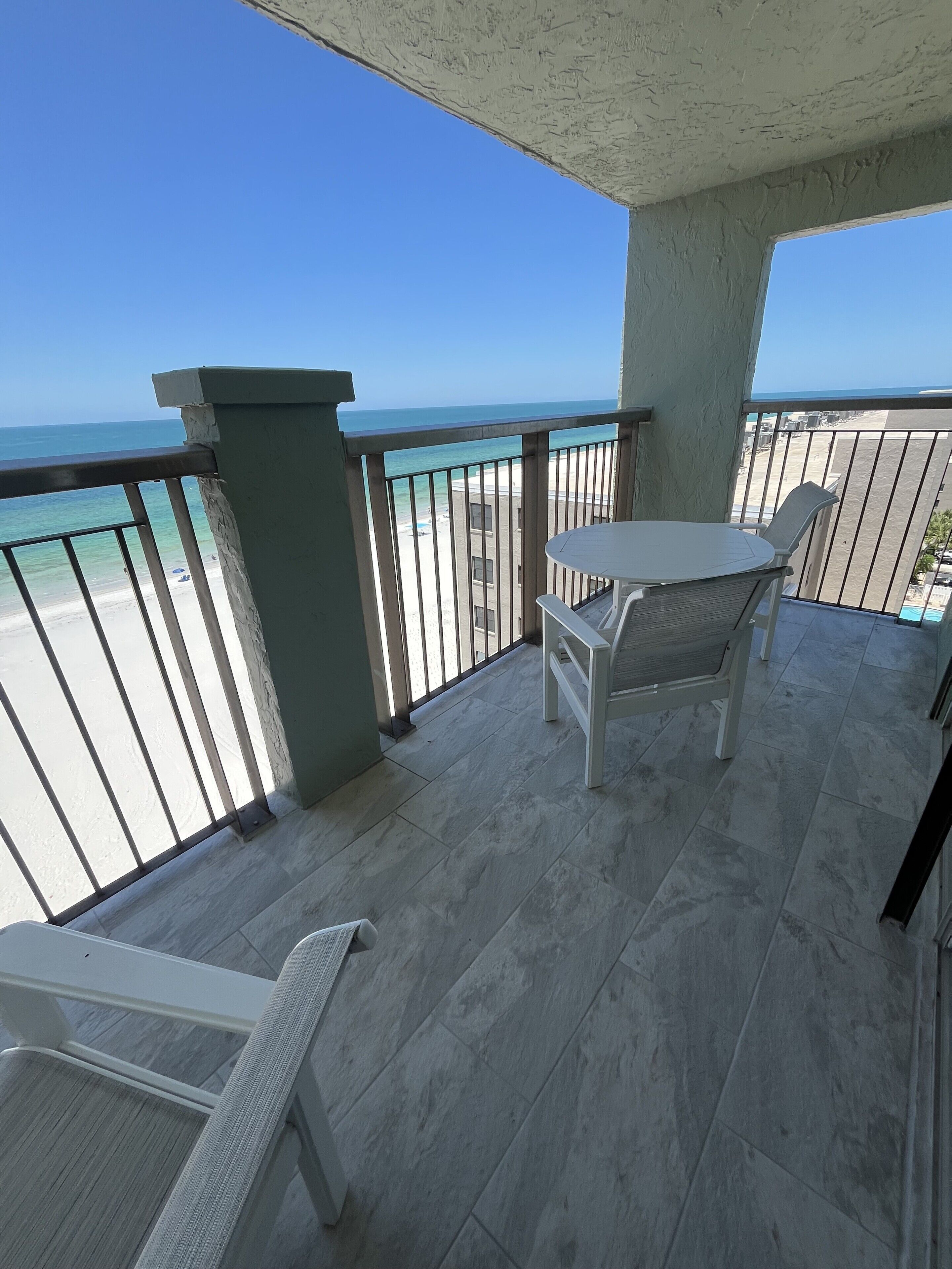A balcony with a view of the beach and ocean, a table, and two chairs.