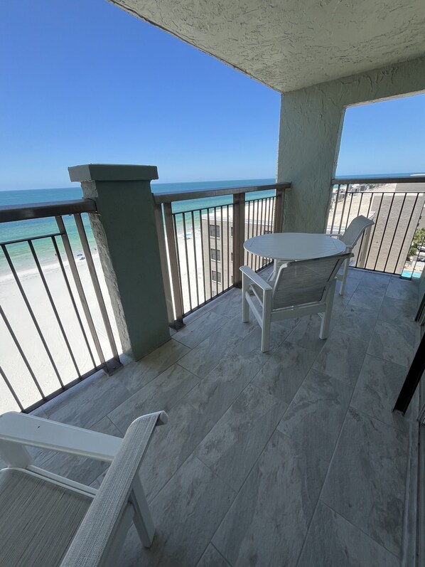 A balcony with a view of the beach and ocean, a table, and two chairs.