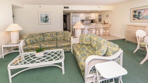 Una sala de estar con alfombra verde, mobiliario de mimbre blanco y una cocina al fondo.