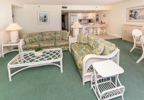 A living room with a green carpet, white wicker furniture, and a kitchen in the background.
