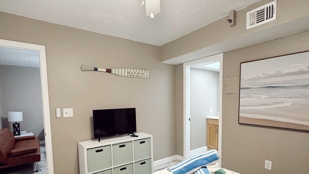 A bedroom with a bed, a television, a ceiling fan, and a beach-themed wall decoration.